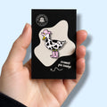 Hand holding a card with an enamel pin badge of a goose with cow print feathers wearing a pink cowboy hat and boots.