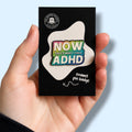 Enamel pin badge packaging with colorful text 'NOW THAT'S WHAT I CALL ADHD' held in a hand against a light blue background.