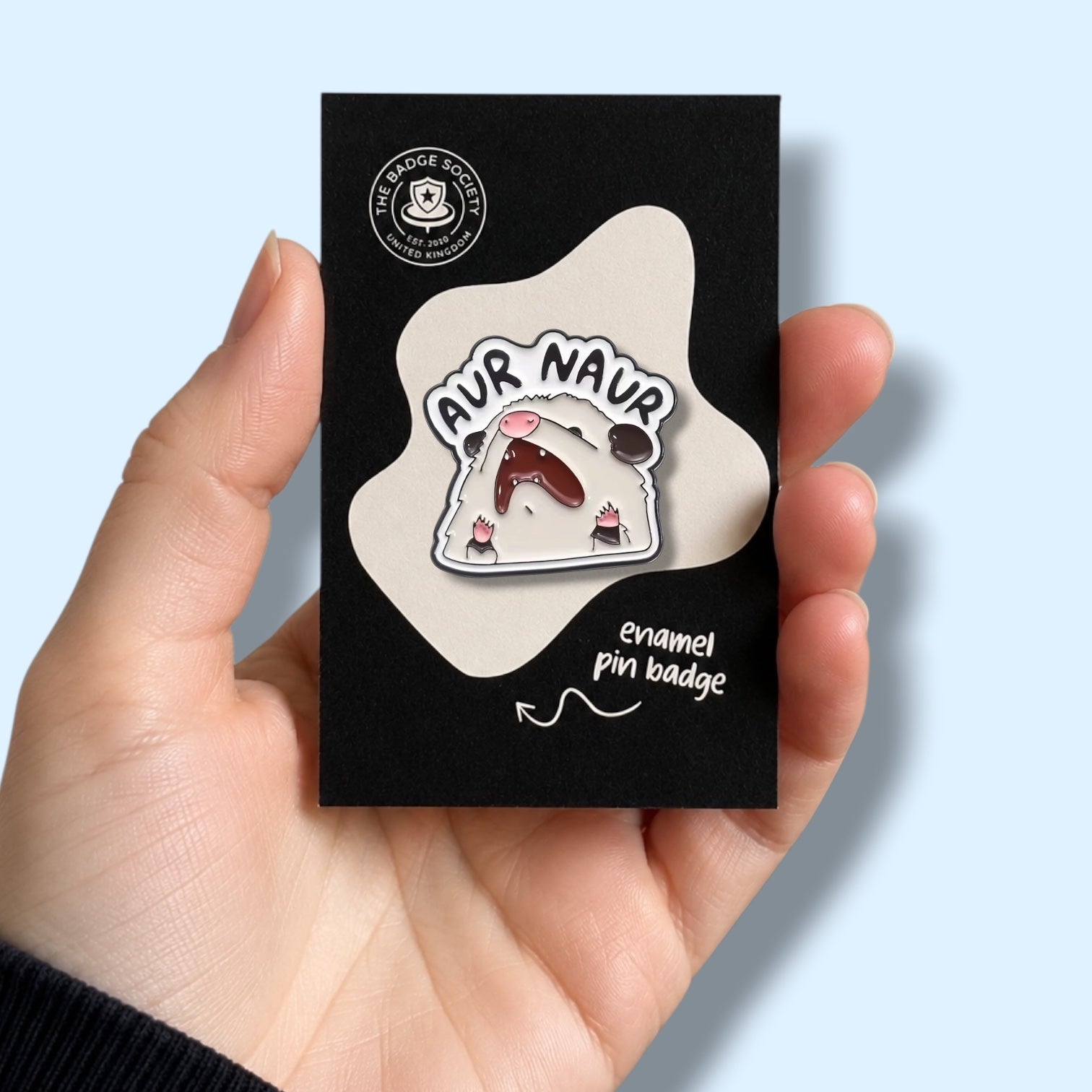 Enamel pin badge on a black card held by a hand against a light blue background. The badge is a screaming opossum with the words Aur Naur. 