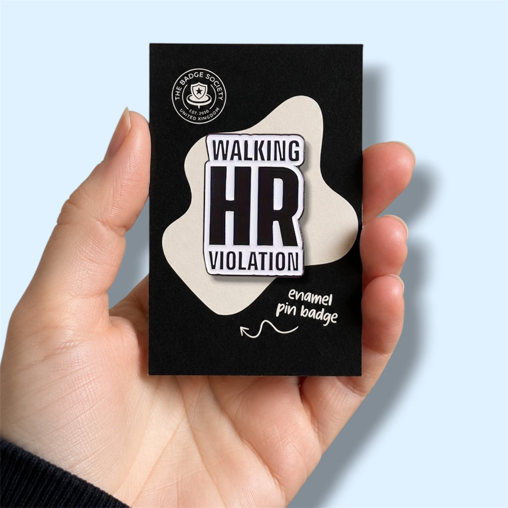 Enamel pin badge packaging with 'Walking HR Violation' text on enamel pin held in a hand against a light blue background.