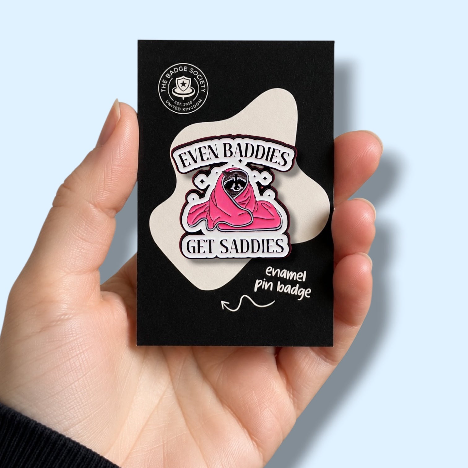 Hand holding a black card with an enamel pin badge featuring a raccoon wearing a blanket with the phrase even baddies get baddies.