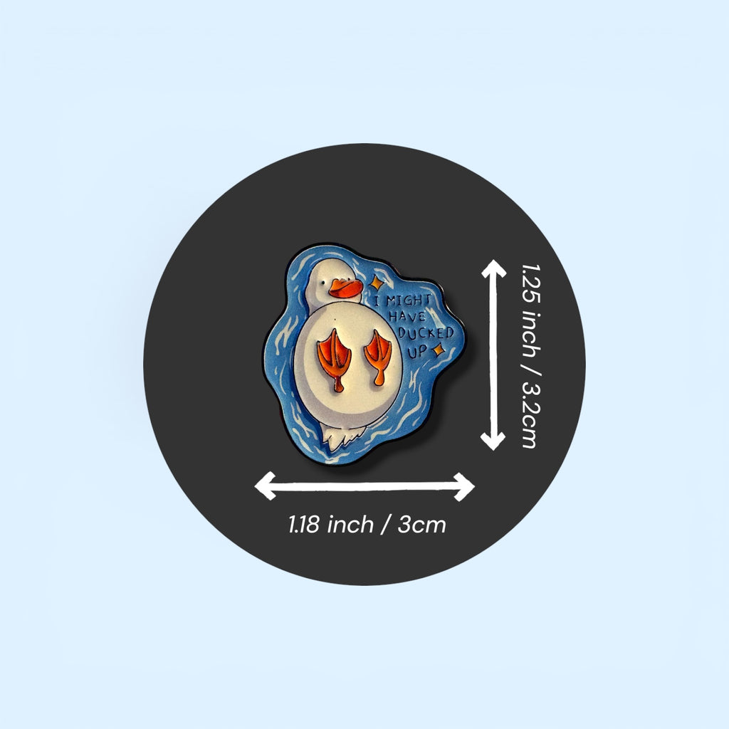 Pin with measurements on a black circle against a light blue background. Enamel pin badge on the black circle of a duck swimming on it's back with the text 'I might have ducked up' 