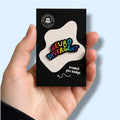Hand holding a card with a colorful 'Neuro Divergent' enamel pin badge against a light blue background.