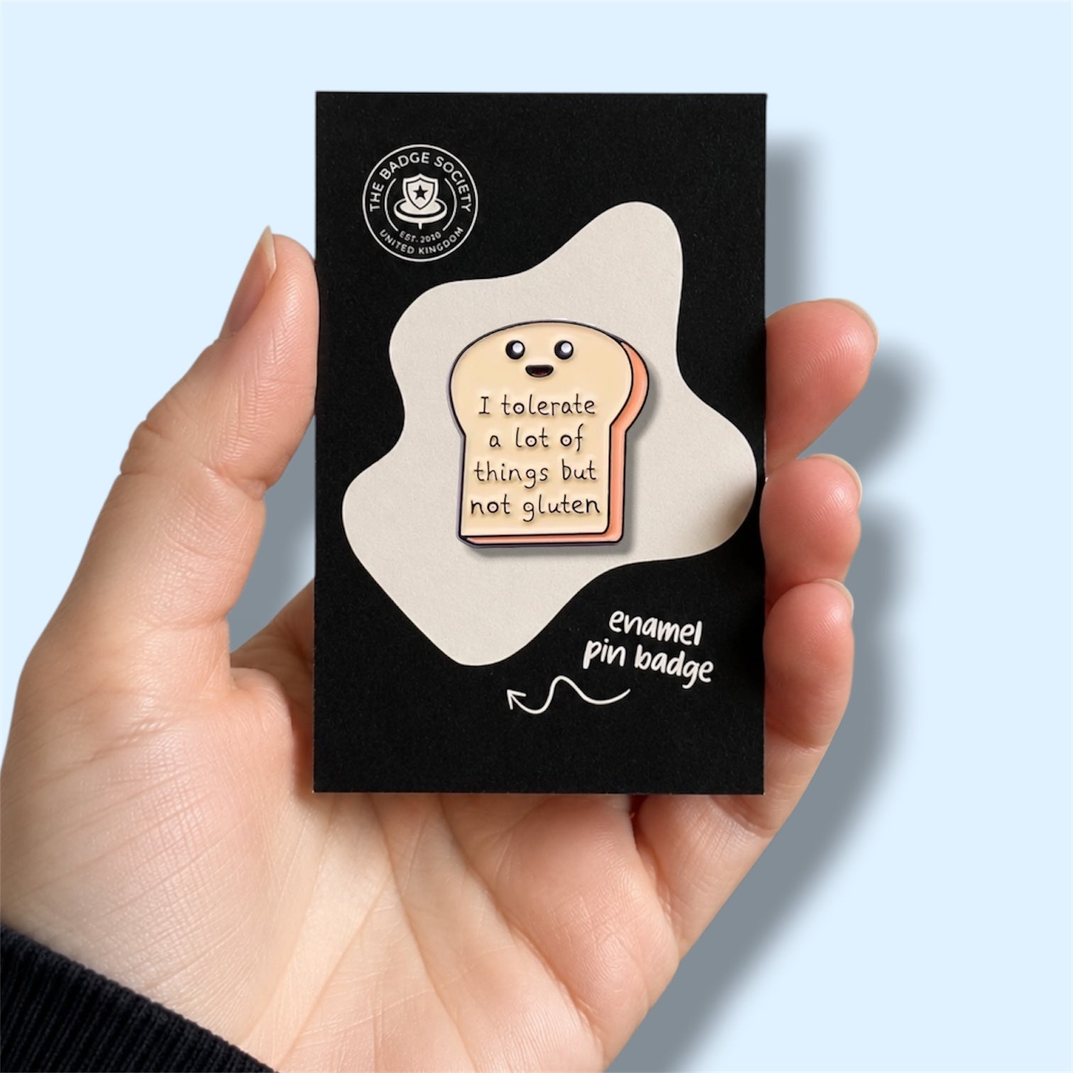 Enamel pin badge packaging held by a hand with a light blue background. Enamel pin is a piece of bread with text saying 'I tolerate a lot of things but not gluten' 