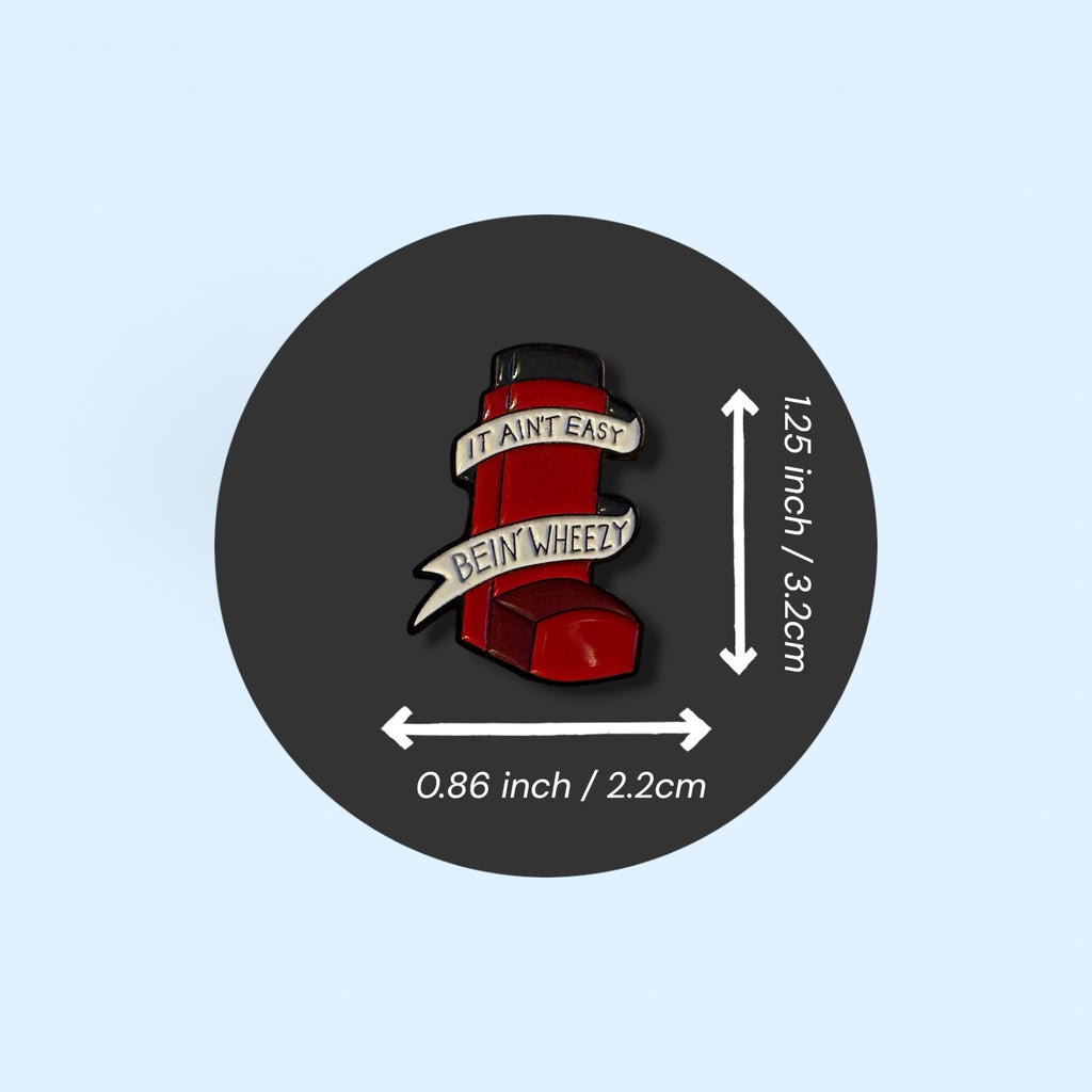 Red and black pin with text on a dark gray circle with measurements on a light blue background. Featuring an enamel pin as a red asthma inhaler design with the text 'it ain't easy being wheezy'.