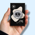 Enamel pin badge in packaging held by a hand against a light blue background. Enamel pin badge is a dog's nose with a heart wreath style surrounding it with the words 'snoot-booper club' 