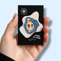Enamel pin badge in packaging held by a hand against a light blue background. The enamel pin is a duck swimming on it's back with the text 'I might have ducked up'.