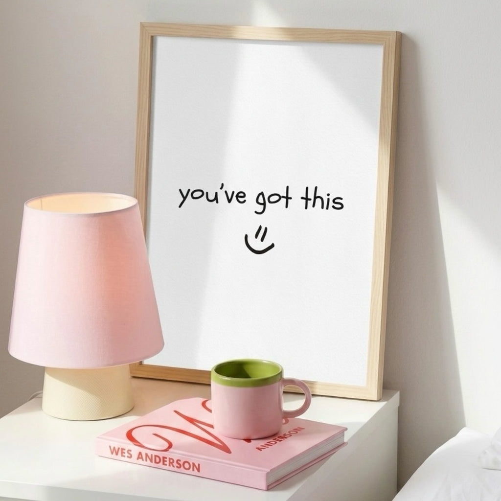 A light wooden framed print sits on a bedside or tabletop surface, displaying the handwritten message “you’ve got this :)” in simple black text. To the left is a pink table lamp with a white base, softly illuminating the scene. In front of the lamp rests a pink book titled WES ANDERSON, with a pink mug featuring a green interior placed neatly on top. The arrangement creates a warm, calm, and encouraging atmosphere.