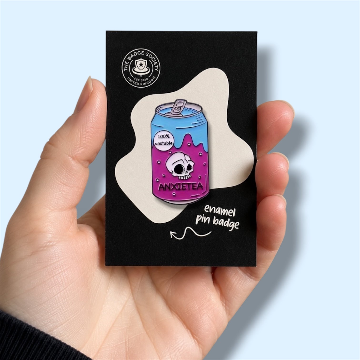 Enamel pin badge of a purple can with a skull design and the text 'anxietea' and '100% unstable' on a black card held by a hand. 