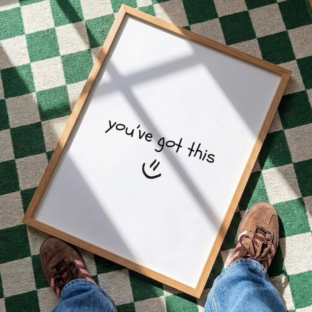 A wooden‑framed print lies flat on a green‑and‑white checkered floor, illuminated by angled natural light that casts soft shadows across its surface. The print shows the handwritten message “you’ve got this :)” in simple black lettering. At the bottom edge of the scene, the lower legs and feet of a person wearing blue jeans and brown shoes are visible, positioned as if they are standing just in front of the print and looking down at it.