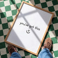 A wooden‑framed print lies flat on a green‑and‑white checkered floor, illuminated by angled natural light that casts soft shadows across its surface. The print shows the handwritten message “you’ve got this :)” in simple black lettering. At the bottom edge of the scene, the lower legs and feet of a person wearing blue jeans and brown shoes are visible, positioned as if they are standing just in front of the print and looking down at it.