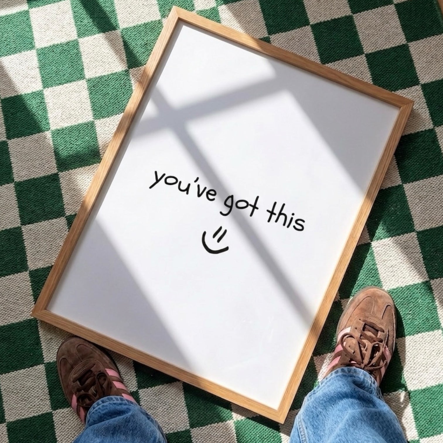 A wooden‑framed print lies flat on a green‑and‑white checkered floor, illuminated by angled natural light that casts soft shadows across its surface. The print shows the handwritten message “you’ve got this :)” in simple black lettering. At the bottom edge of the scene, the lower legs and feet of a person wearing blue jeans and brown shoes are visible, positioned as if they are standing just in front of the print and looking down at it.