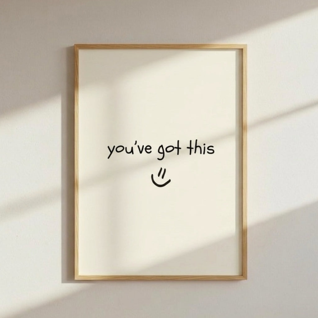 A light wooden frame hangs on a softly sunlit wall, with gentle shadows falling across its surface. Inside the frame is a minimalist white print featuring the handwritten message “you’ve got this :)” in simple black text. The warm lighting and clean design create a calm, encouraging atmosphere.